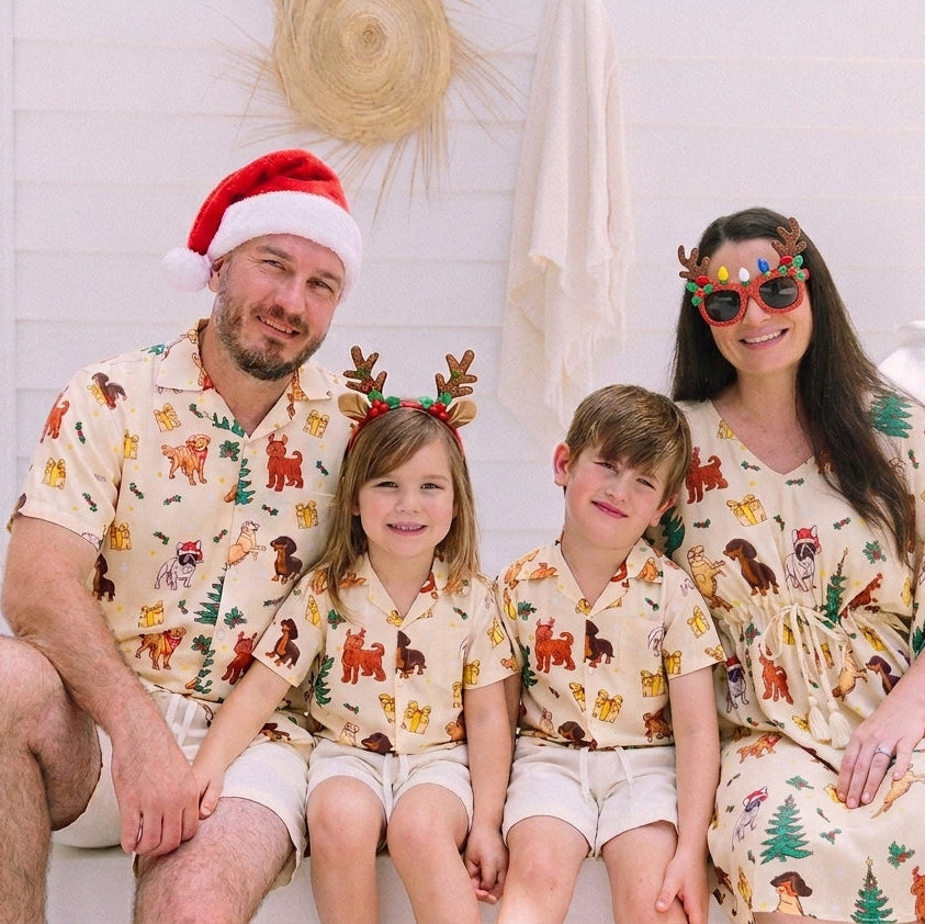 A family of four wearing matching Christmas-themed safari print robes and matching hats, sitting together on a white bench in a bright, modern indoor setting.