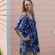 A woman wearing a navy blue and white toile-print kaftan dress, standing outdoors against a peach-colored house siding.