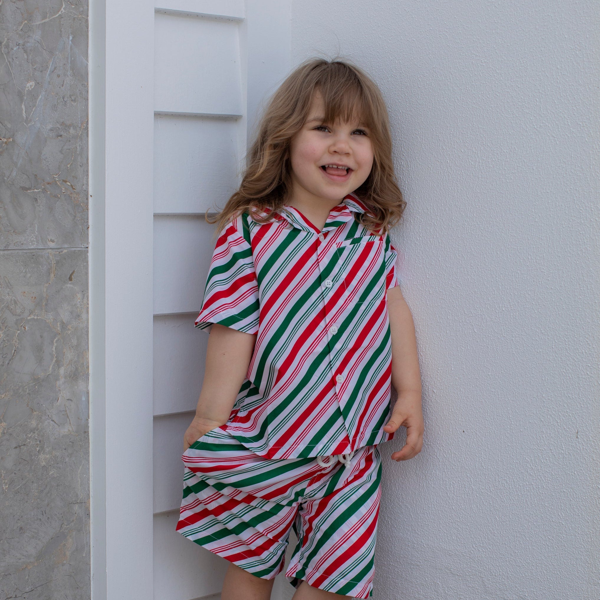 A young girl with blonde hair stands barefoot against a white wall, wearing a matching short-sleeve collared shirt and shorts set with diagonal red, green, and white stripes.