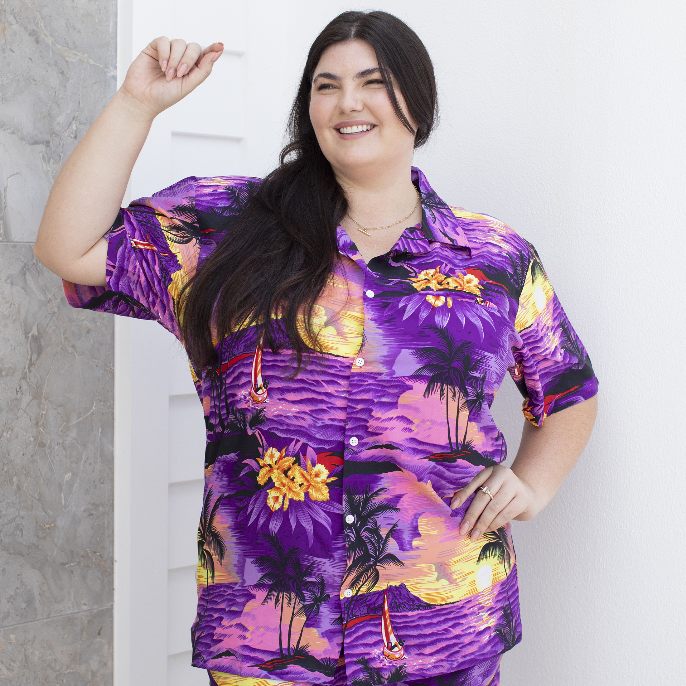 Adult female model wearing a purple Hawaiian shirt and shorts with a tropical design, posing with one hand extended.