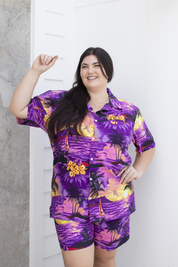 Adult female model wearing a purple Hawaiian shirt and shorts with a tropical design, posing with one hand extended.