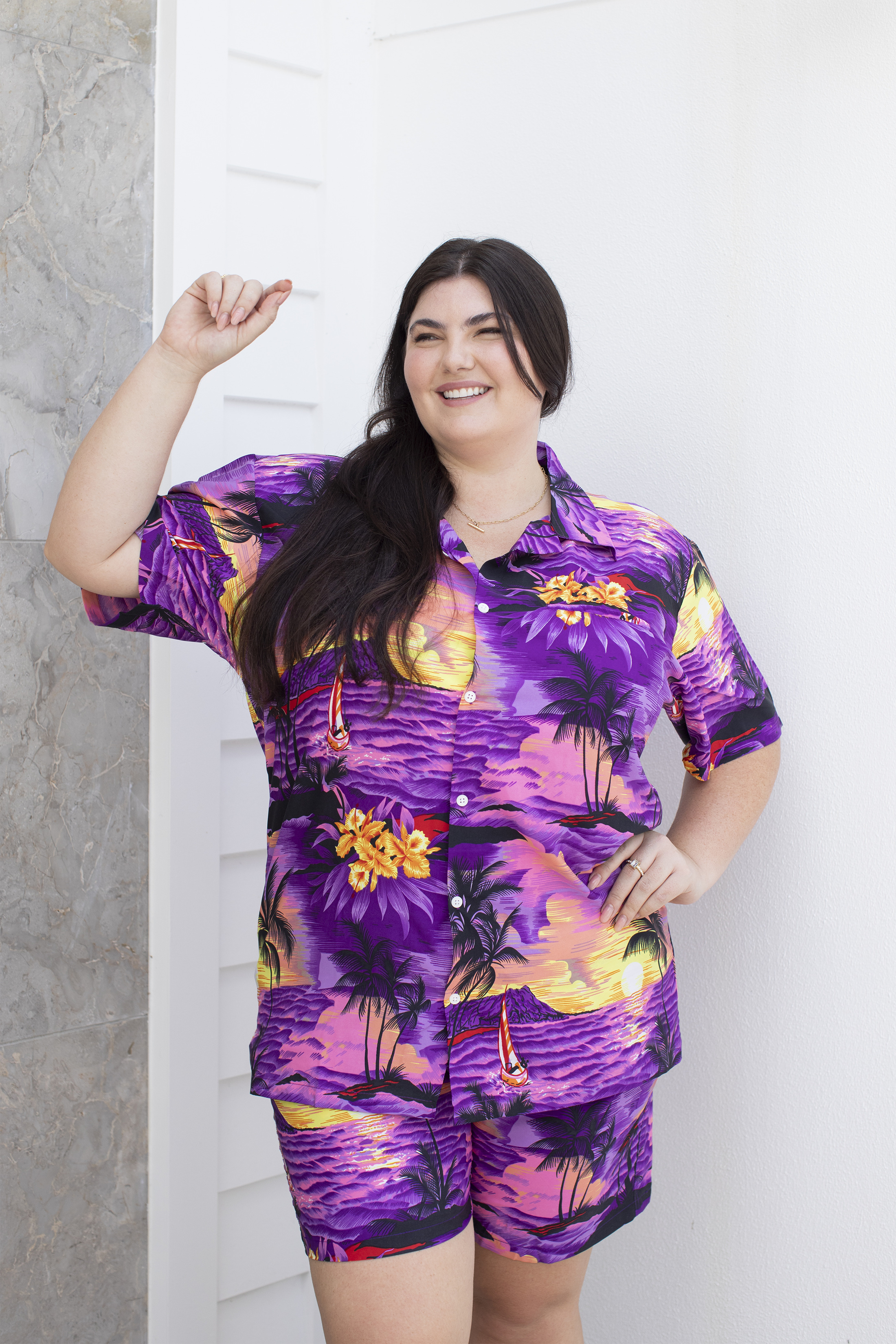 Adult female model wearing a purple Hawaiian shirt and shorts with a tropical design, posing with one hand extended.