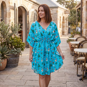 A woman wearing a turquoise blue kaftan dress with a Christmas print of snowmen, Christmas trees, and ornaments, walking in a stone patio courtyard.