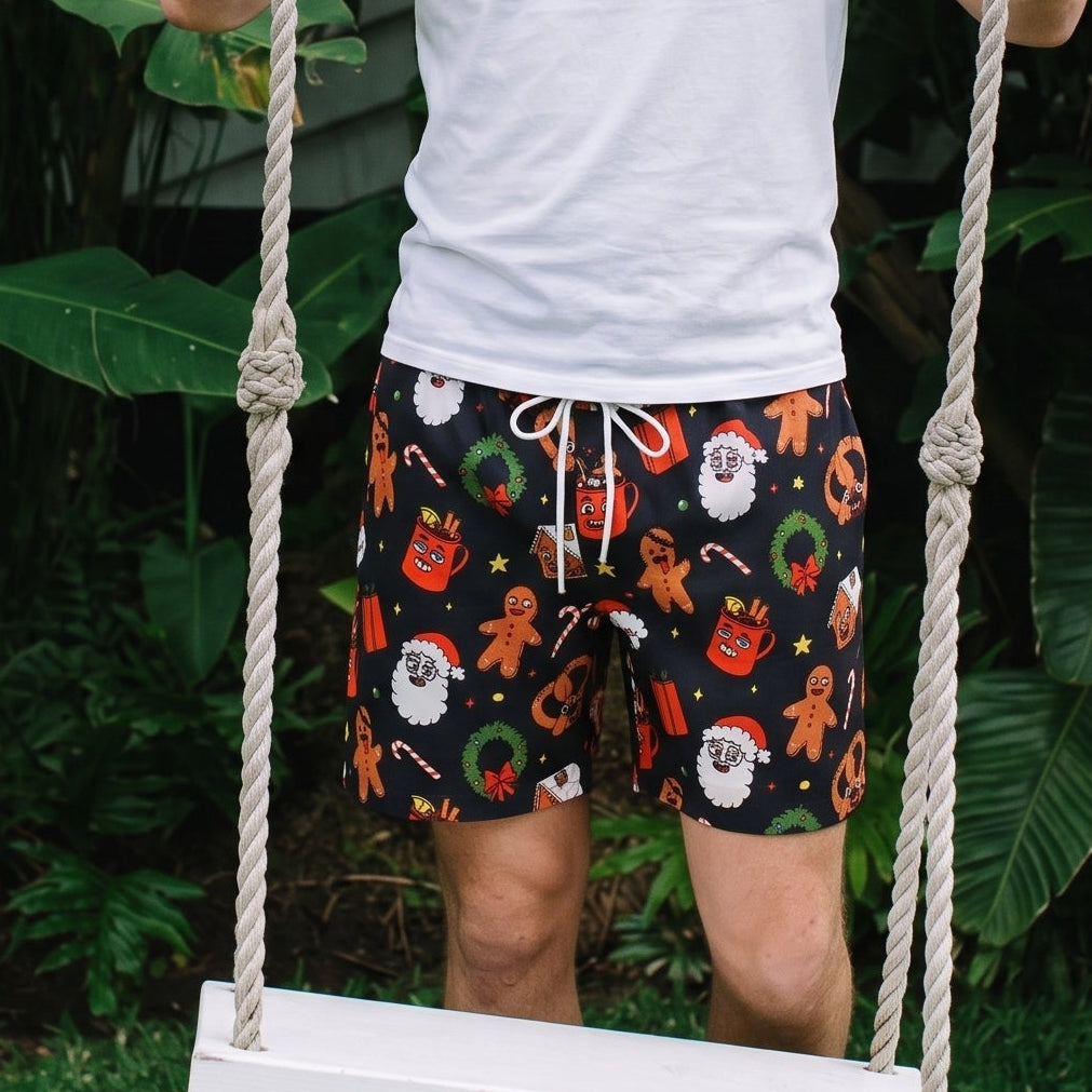 A man stands on a white swing in a lush green outdoor setting, wearing a plain white t-shirt and black swim trunks with a colorful Halloween-themed print of ghosts, pumpkins, and skulls.