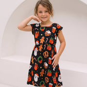 A young girl wearing a black short-sleeve A-line dress with a festive Christmas print of gingerbread men, Santa Claus, candy canes, and ornaments, posing in front of a white arched wall.