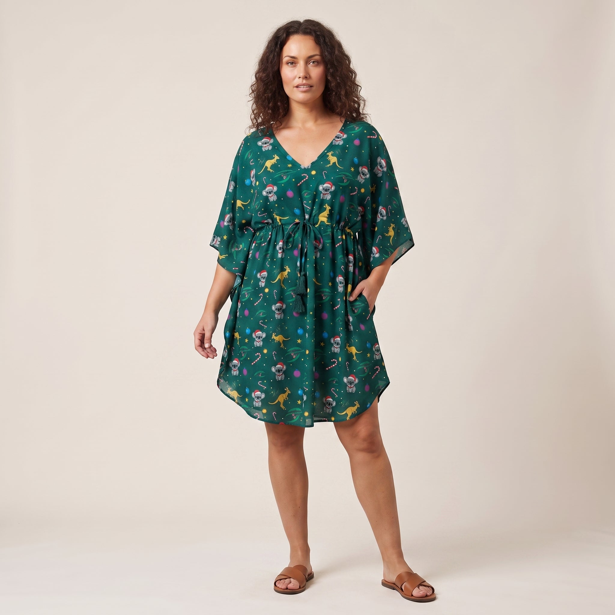 A woman modeling a green kaftan-style dress with a colorful floral and bird print, standing against a plain beige studio background.
