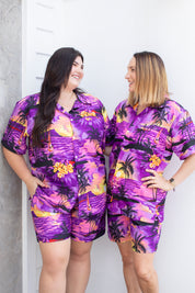 Two women wearing matching purple tropical outfits standing against a white wall.