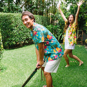 A man and a woman playfully pose on a lush green lawn, both wearing matching vibrant tropical Hawaiian shirts. The man in the foreground is smiling and holding a large machete, while the woman in the background celebrates with her arms raised.