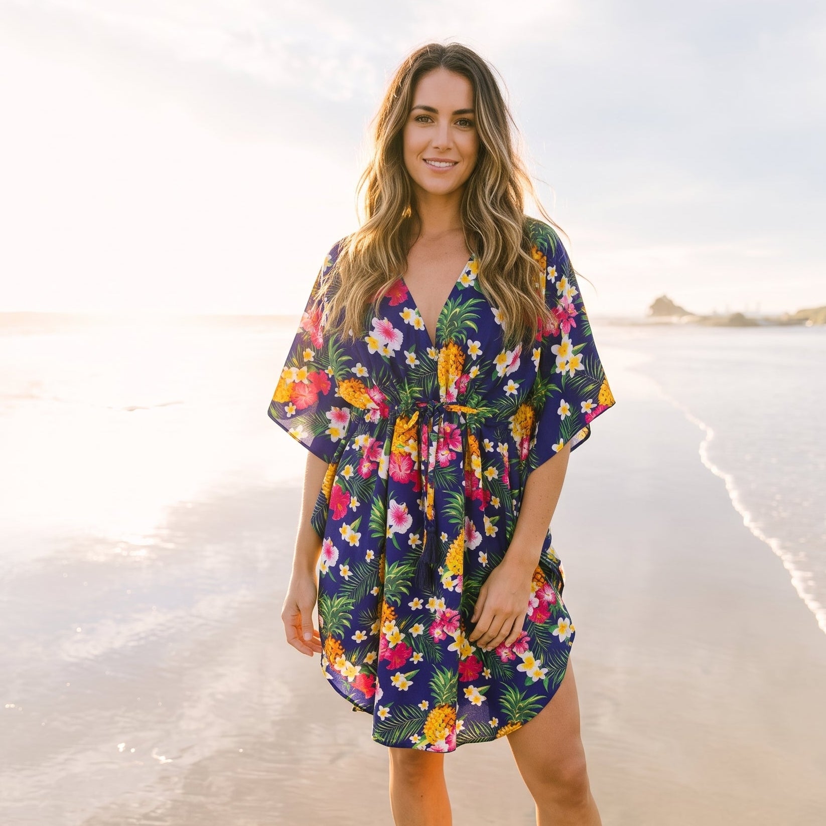 A woman stands barefoot on a sandy beach wearing a short, loose floral dress with wide sleeves and a tie waist. The dress has a navy base with a colorful tropical print including pink, yellow, green, and white flowers.