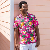 A man wearing a bright pink short-sleeve button-up shirt with a tropical pineapple and white flower print, paired with white pants, standing against a peach-colored house exterior.
