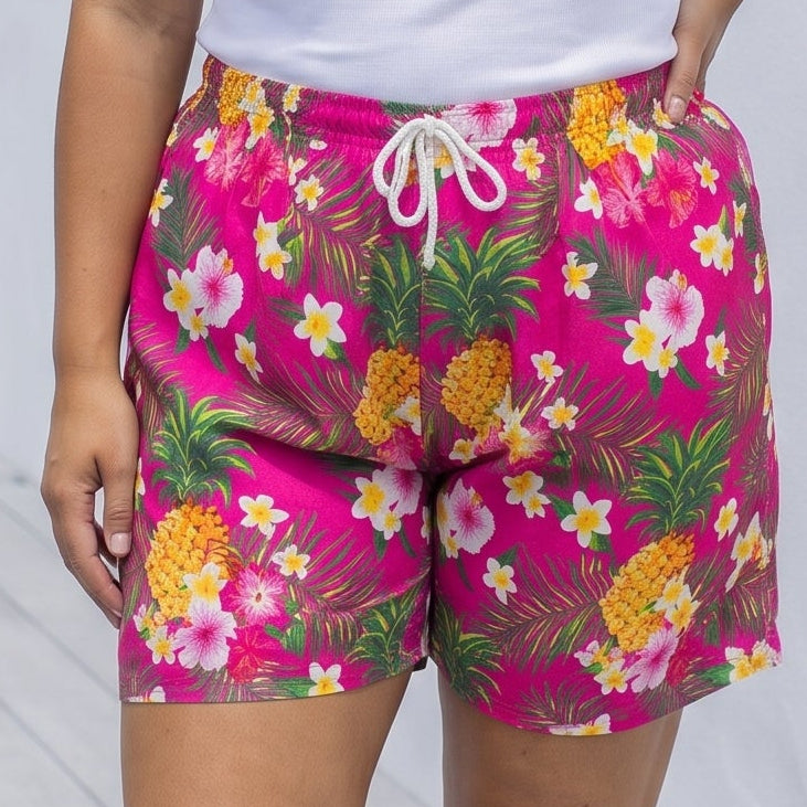 A woman modeling bright pink floral shorts with a white tank top, standing on a light wooden deck against a white wall.