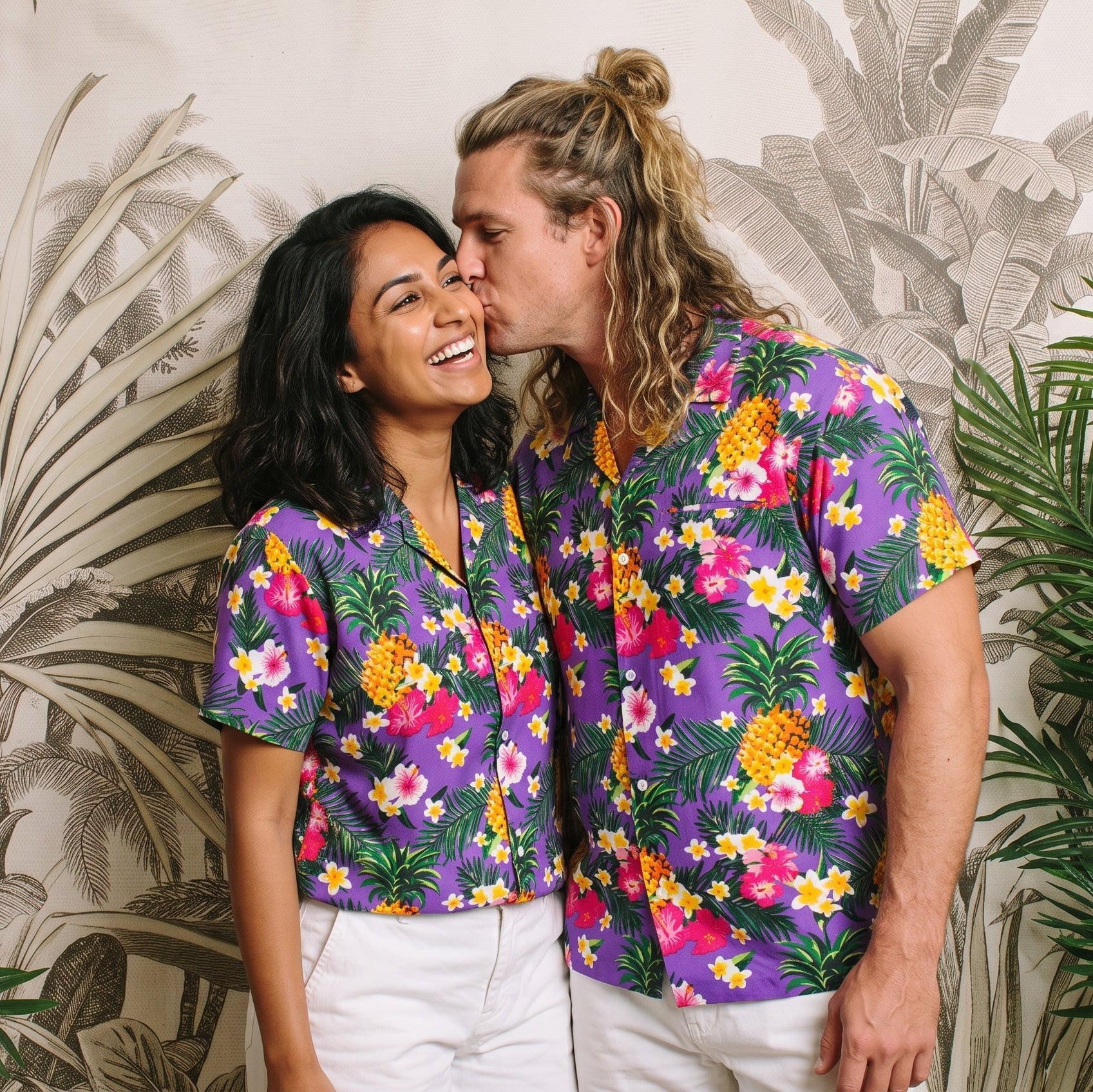 A couple wearing matching purple short-sleeve button-up shirts with a colorful tropical floral and pineapple print, standing in front of a tropical palm leaf wallpaper background with green plants.