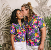 A couple wearing matching purple short-sleeve button-up shirts with a colorful tropical floral and pineapple print, standing in front of a tropical palm leaf wallpaper background with green plants.