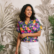 A woman wearing a short-sleeve button-up Hawaiian shirt with a colorful tropical floral and pineapple print on a purple background, paired with white pants, standing in front of a tropical palm leaf patterned wall.