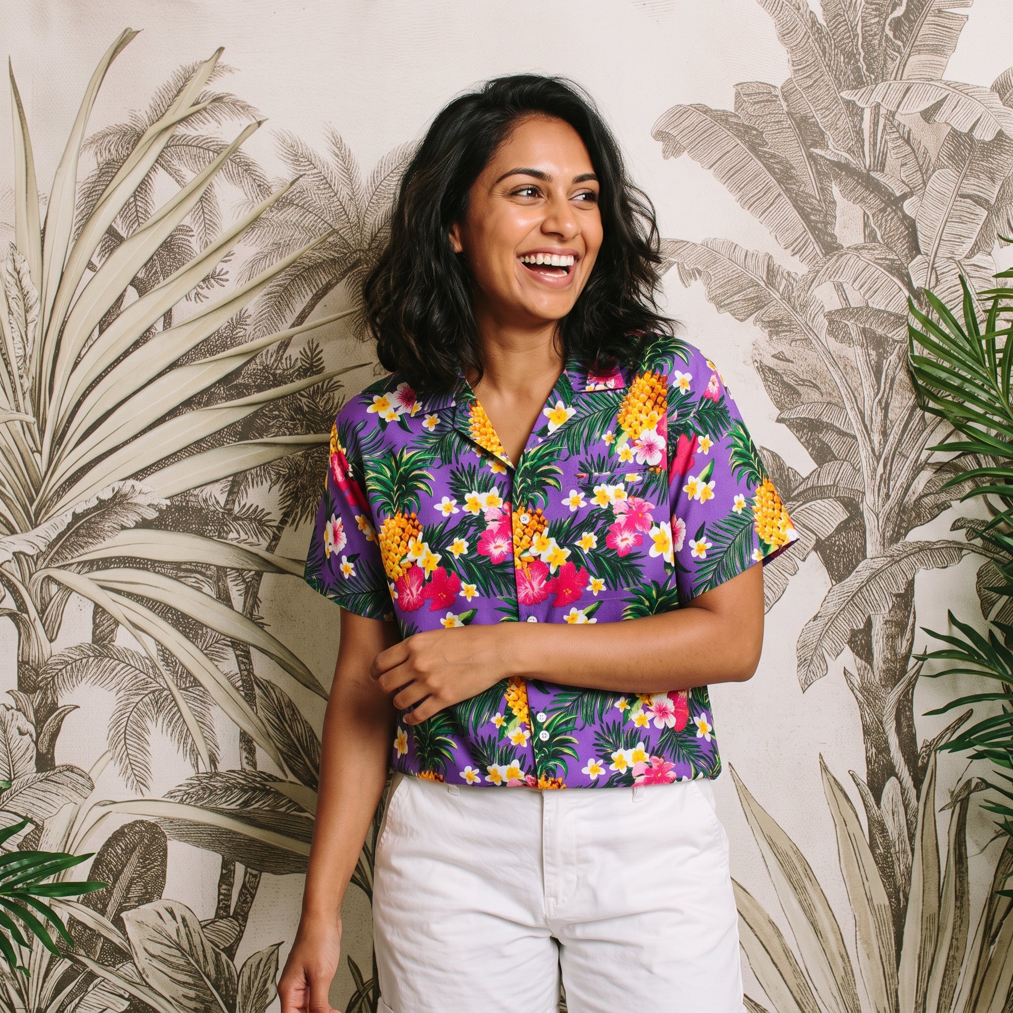 A woman wearing a short-sleeve button-up Hawaiian shirt with a colorful tropical floral and pineapple print on a purple background, paired with white pants, standing in front of a tropical palm leaf patterned wall.