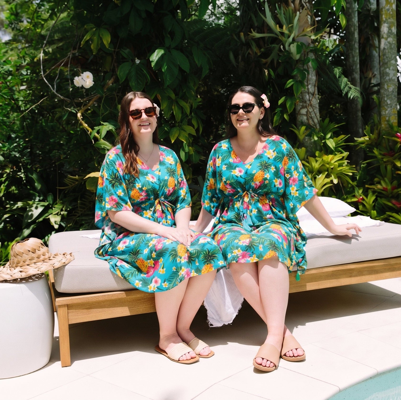 Two women sitting side by side on a wooden lounge daybed outdoors by a pool, wearing matching teal and orange floral kimono-style dresses. Lush tropical greenery surrounds the patio setting.