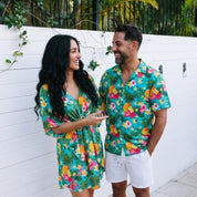 A man and a woman standing outdoors in front of a white fence, both wearing matching tropical floral print short-sleeve shirts and a matching women's short wrap dress. The print features green, pink, yellow, and blue flowers with palm leaves.