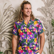 A man wearing a purple short-sleeve button-up shirt with a colorful tropical floral and pineapple print, paired with white shorts, standing in front of a tropical leaf patterned wall.
