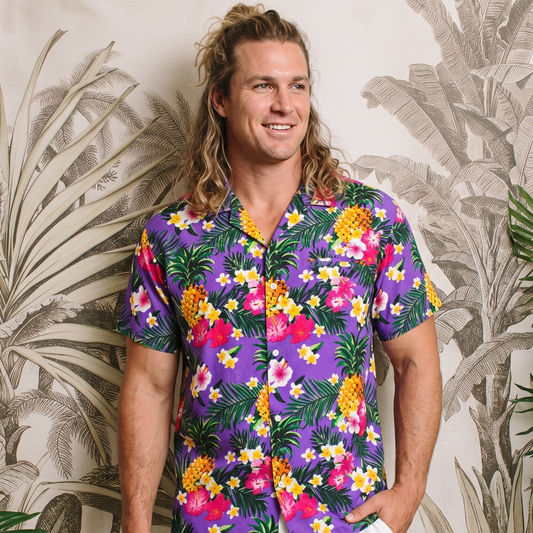 A man wearing a purple short-sleeve button-up shirt with a colorful tropical floral and pineapple print, paired with white shorts, standing in front of a tropical leaf patterned wall.
