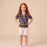A young girl with red hair stands against a solid beige studio background, wearing a navy blue short-sleeve button-up shirt with a colorful fast-food print (burgers, hot dogs, pizza, fries) and cream shorts.