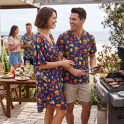 A man and woman standing together at an outdoor patio grill, both wearing matching navy blue short-sleeve button-up shirts covered in a colorful hamburger print. The woman wears a knee-length matching dress, and the man wears khaki shorts.