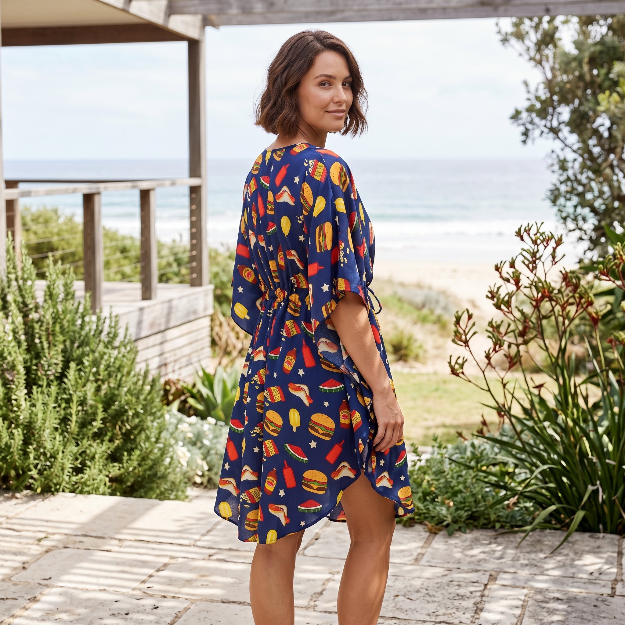 A woman wearing a navy blue dress with a colorful beach-themed print of palm trees, sunglasses, and starfish, standing on a stone patio with a beach and ocean background.