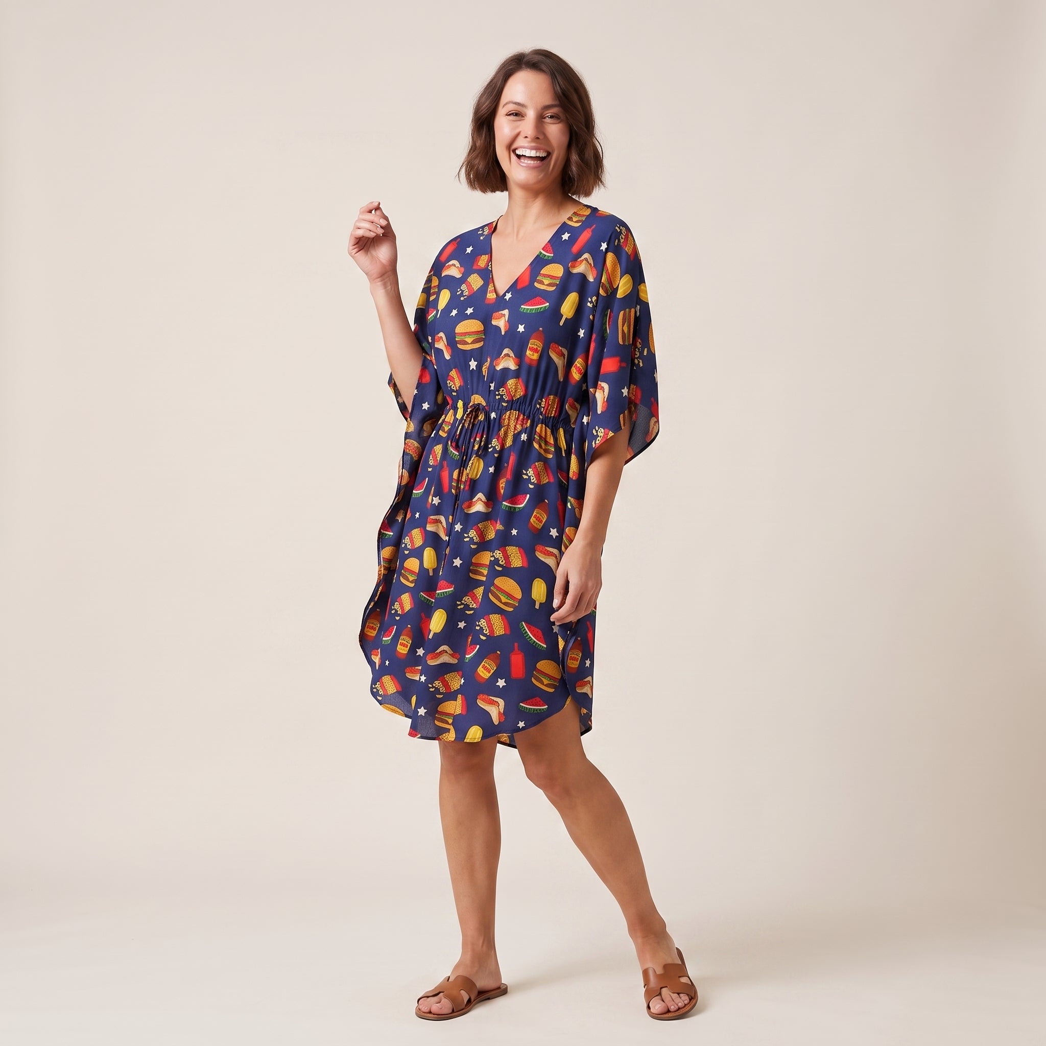 A woman modeling a navy blue kaftan dress with a colorful burger print, standing against a plain beige studio background.