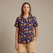 A woman modeling a navy blue short-sleeve button-up shirt with a colorful fast-food print, paired with beige shorts, against a solid beige studio background.