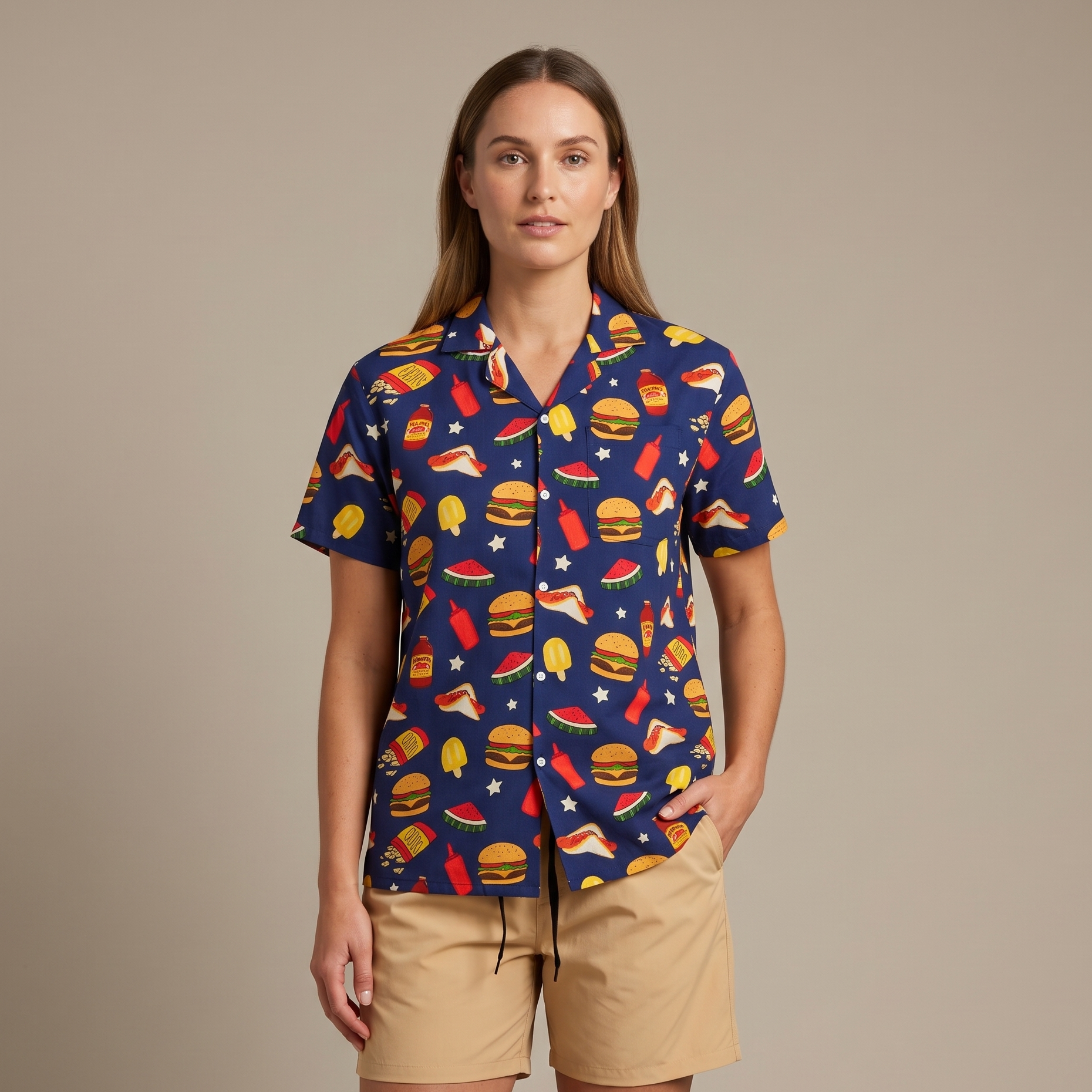 A woman modeling a navy blue short-sleeve button-up shirt with a colorful fast-food print, paired with beige shorts, against a solid beige studio background.