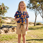 A young boy stands outdoors on a sandy path in a park-like setting, wearing a short-sleeve button-up shirt with a colorful tropical print and beige shorts with a black drawstring.