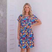 A woman modeling a short-sleeve knee-length dress with a colorful cartoon pattern, standing against a white wall on a wooden deck.