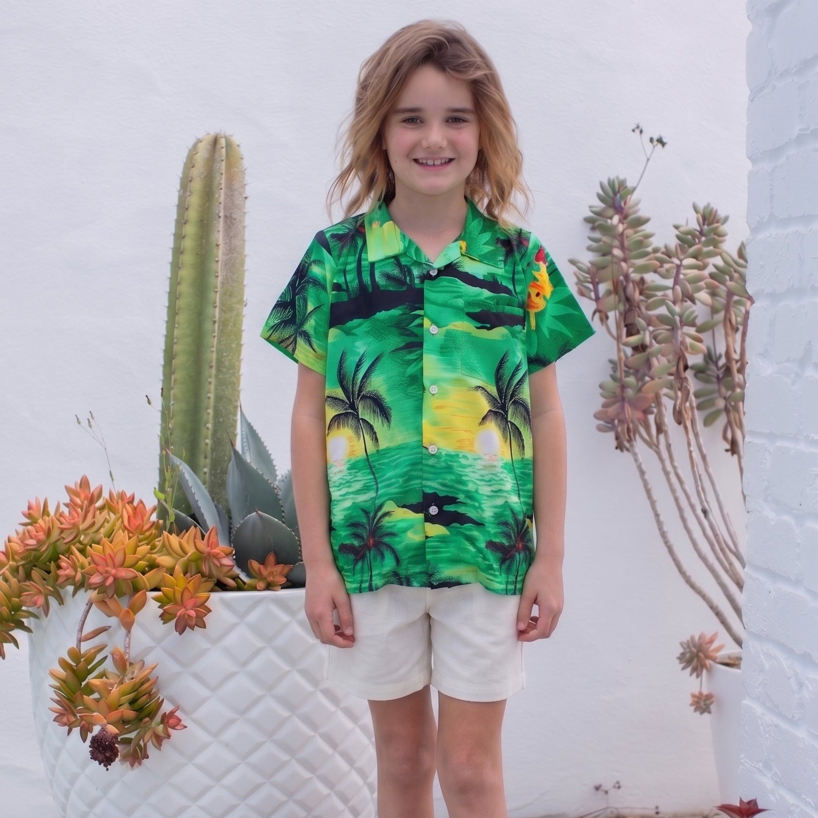 A young boy stands barefoot on a wooden deck wearing a vibrant green tropical print short-sleeve button-up shirt and off-white shorts, set against a white brick wall with potted cacti.