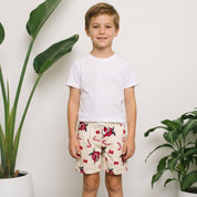 A young boy stands barefoot in a studio setting, wearing a white t-shirt and cream swim trunks with a red and black flamingo pattern. He is framed by two large potted green plants against a white wall.