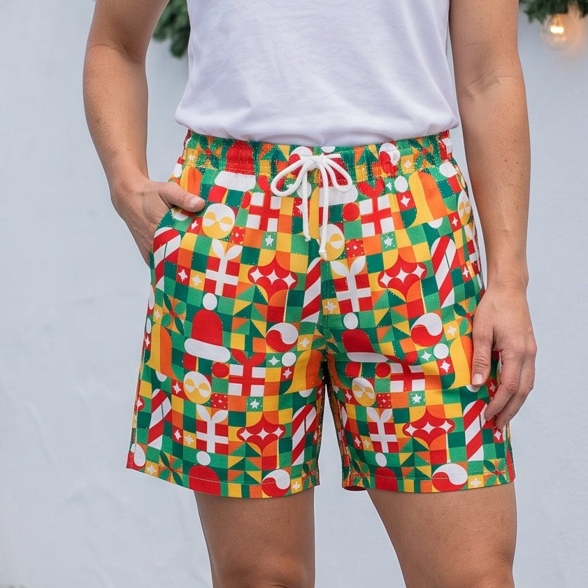 A woman stands on a light gray wooden deck wearing colorful geometric print shorts, a white tank top, and black slide sandals, with a white wall and holiday garlands in the background.
