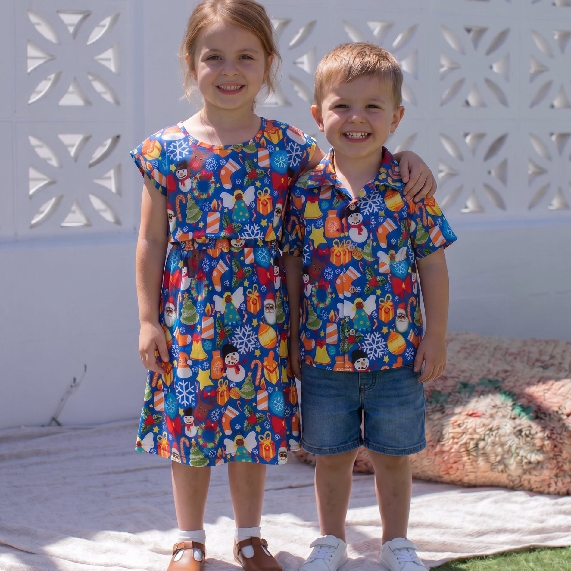 A young girl and boy standing outdoors in front of a white decorative wall. The girl wears a short-sleeve blue dress and the boy wears a matching blue short-sleeve button-up shirt with denim shorts.