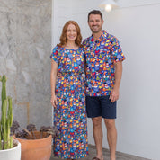 A man and a woman standing together on a porch wearing matching colorful short-sleeve button-up shirts and a matching maxi dress, with the man wearing navy shorts and sandals.