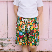 A young girl stands in front of a weathered pink wooden barn door, wearing colorful patterned shorts with a white drawstring, a white t-shirt, and gray slip-on shoes.