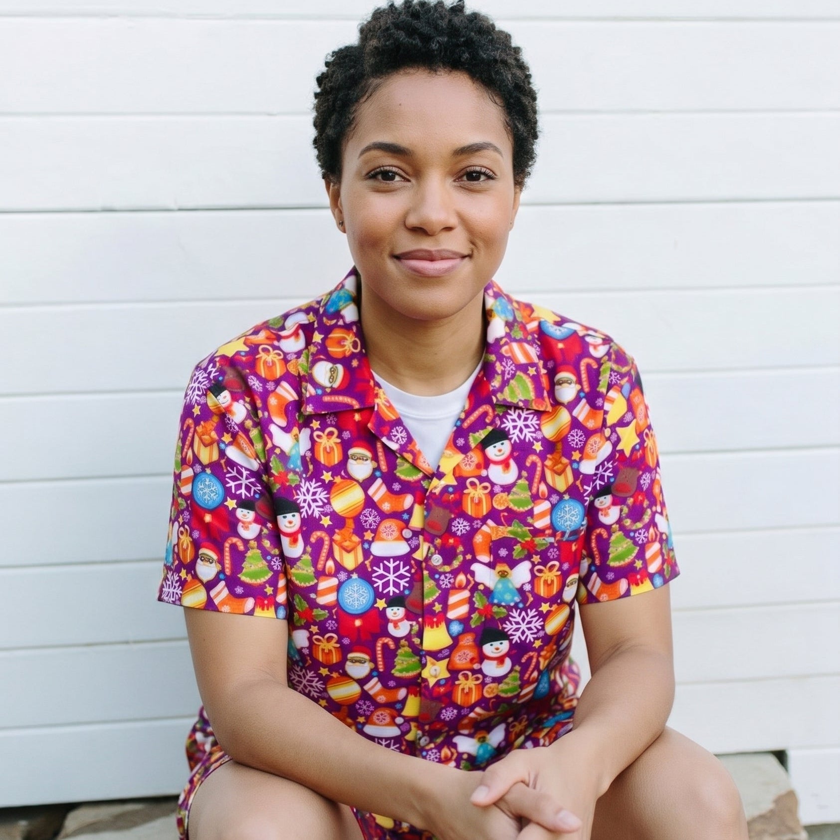 A woman with short dark curly hair sits on stone steps in front of a white shiplap wall, wearing a short-sleeve button-up scrub top and matching shorts with a colorful cartoon pattern on a purple background.