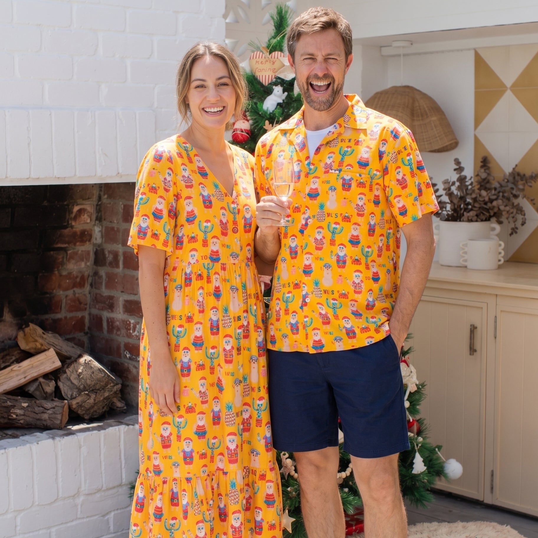 A man and a woman standing together in a cozy indoor setting, wearing matching bright yellow holiday-themed short-sleeve shirts and a dress with a colorful Santa Claus print. The woman holds a glass of champagne, and a Christmas tree with wrapped gifts is visible behind them.