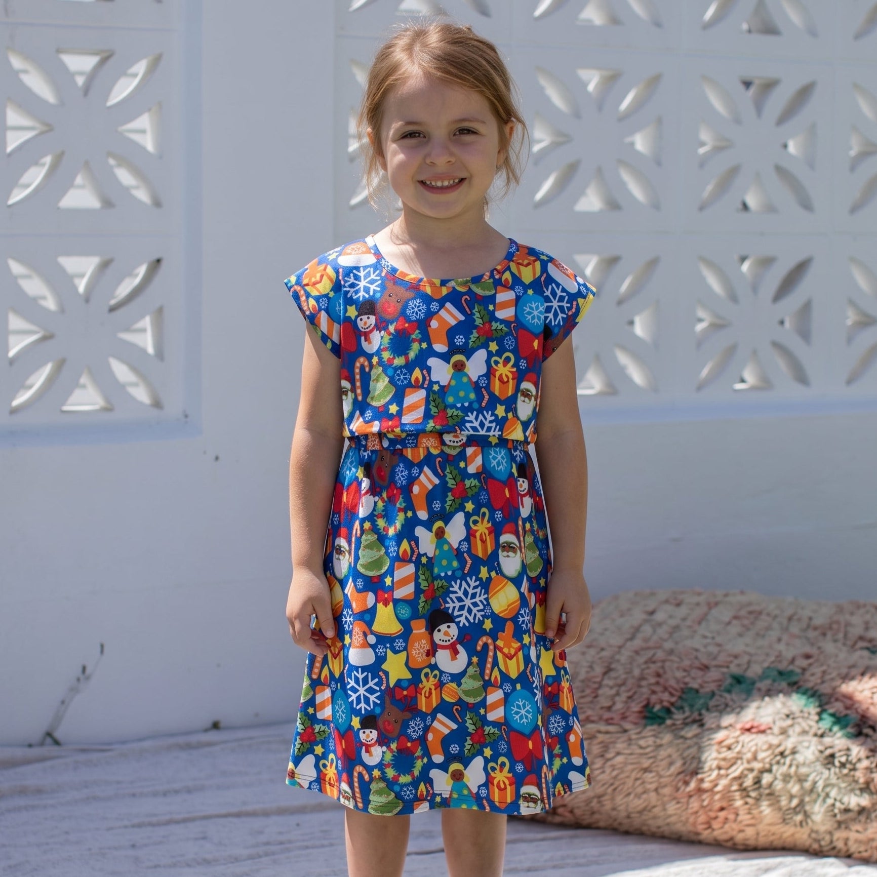 A young girl stands barefoot outdoors wearing a short-sleeve knee-length dress with a colorful holiday-themed print featuring snowflakes, ornaments, and festive illustrations on a blue background. She is standing on a white blanket in front of a white decorative fence.