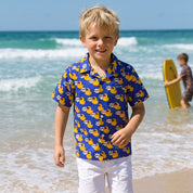 A young blond boy walking on a sandy beach wearing a blue short-sleeve button-up shirt with a yellow rubber duck pattern and white shorts, with ocean waves and a blurred surfer in the background.