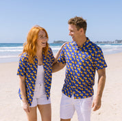 A couple walking on a sandy beach wearing matching blue short-sleeve button-up shirts with a yellow pattern, white shorts, and white tank tops under a clear blue sky.