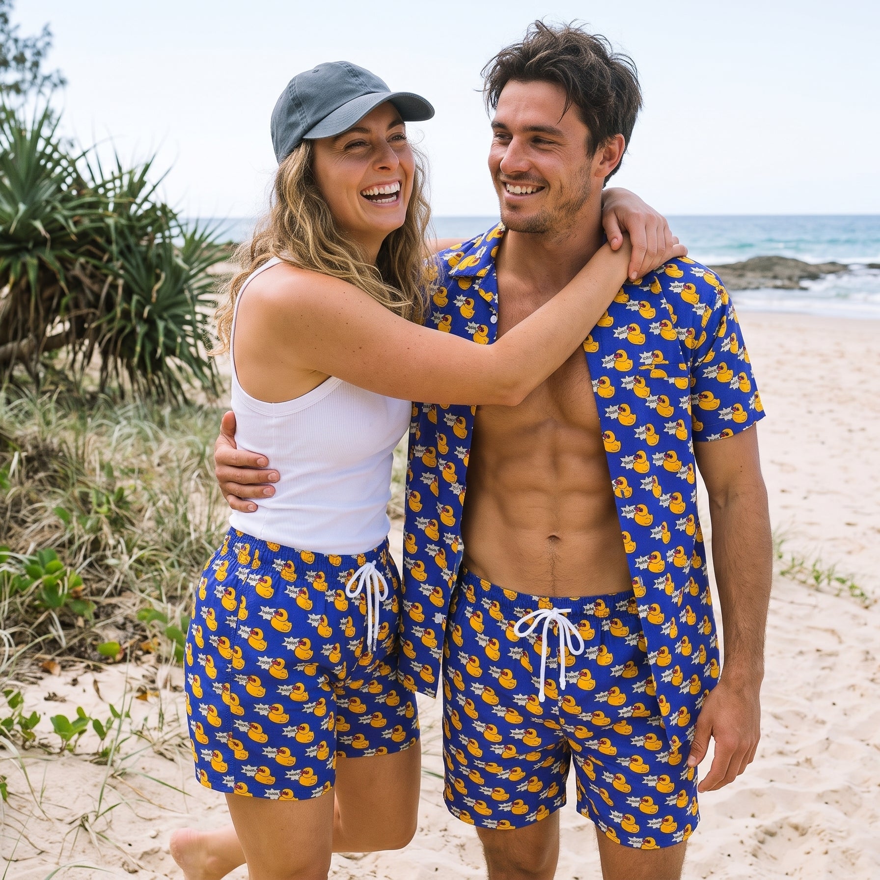 A smiling couple standing on a sandy beach wearing matching blue swim trunks and a matching blue short-sleeve shirt with a yellow duck print. The woman wears a white tank top and a gray cap, and the man is shirtless.