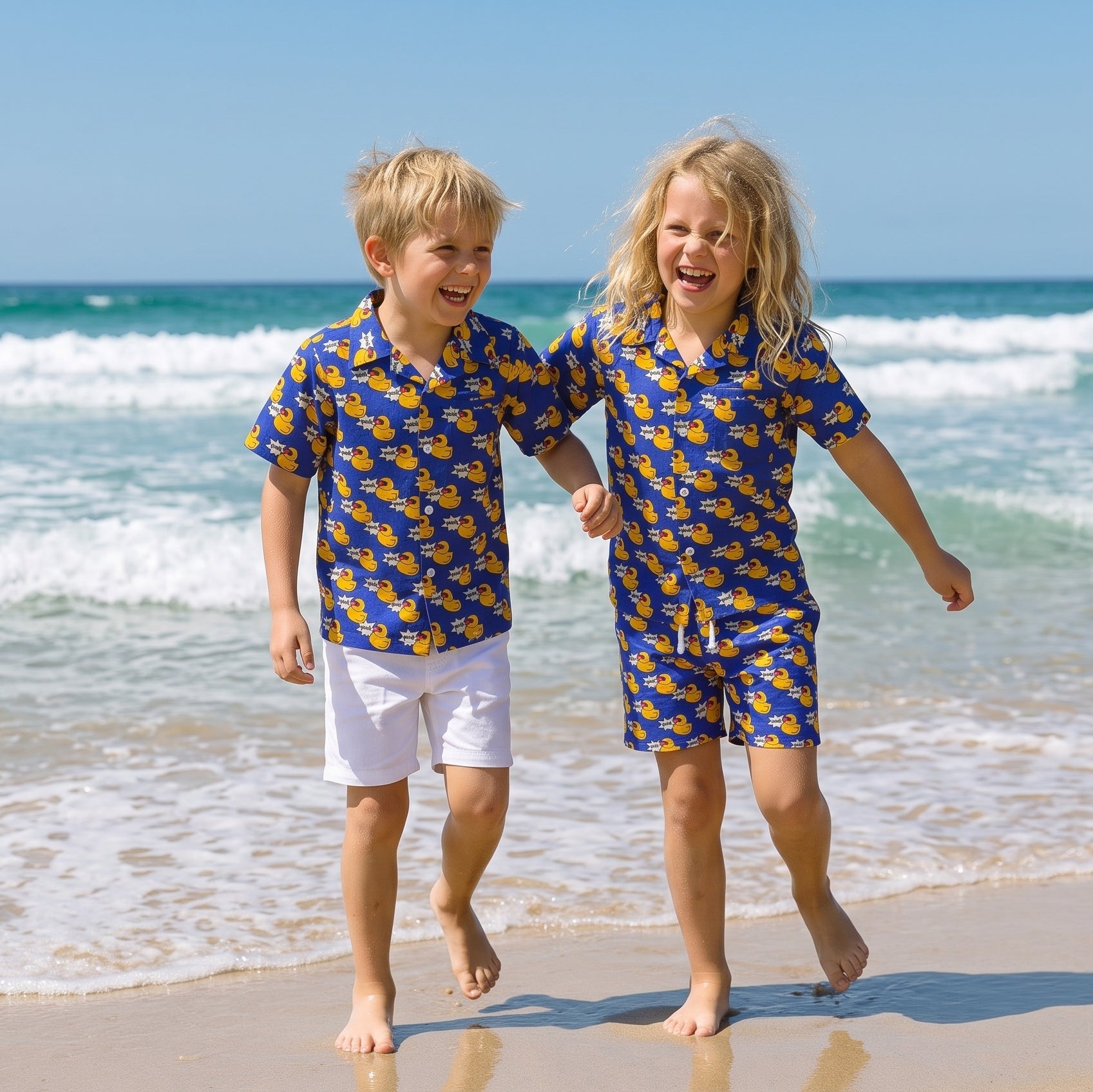 Two young children running and laughing on a sandy beach, wearing matching blue and yellow patterned short-sleeve shirts and shorts sets.