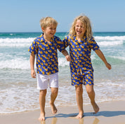 Two young children running and laughing on a sandy beach, wearing matching blue and yellow patterned short-sleeve shirts and shorts sets.