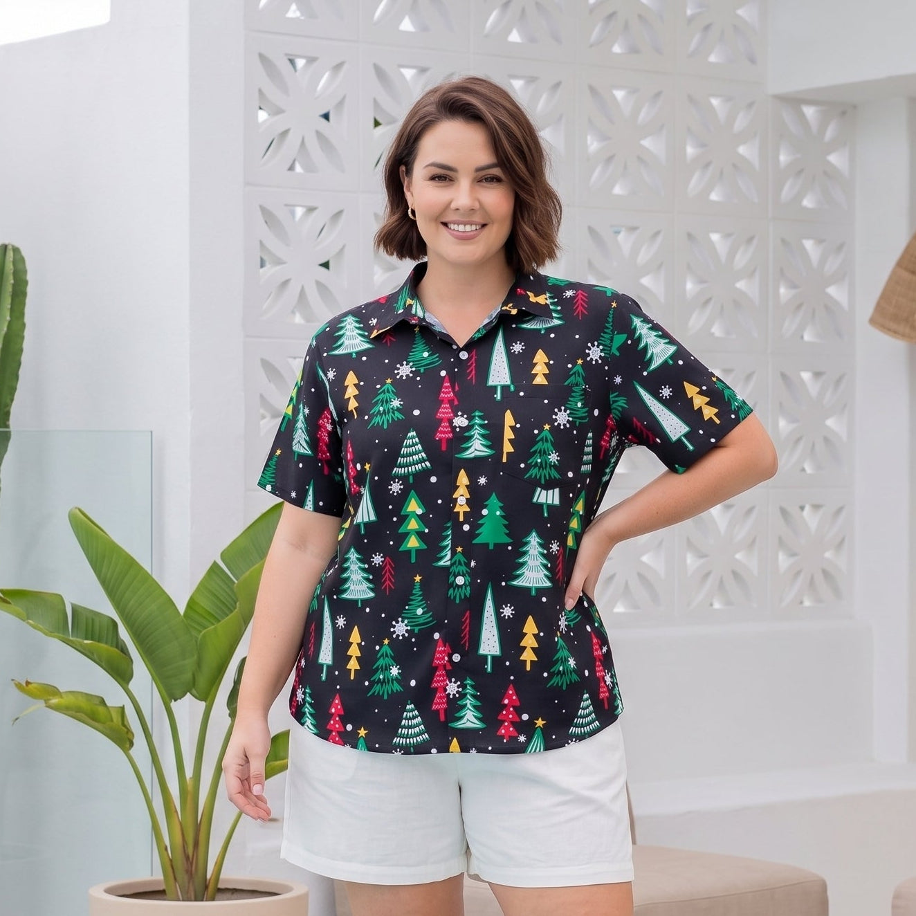 A woman modeling a short-sleeve button-up shirt with a colorful Christmas tree pattern, paired with white shorts, standing on a wooden deck in a bright, modern outdoor patio setting.