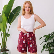 A woman stands indoors wearing a white tank top and red holiday print shorts with a white drawstring. Large green potted plants are visible on both sides against a white wall.