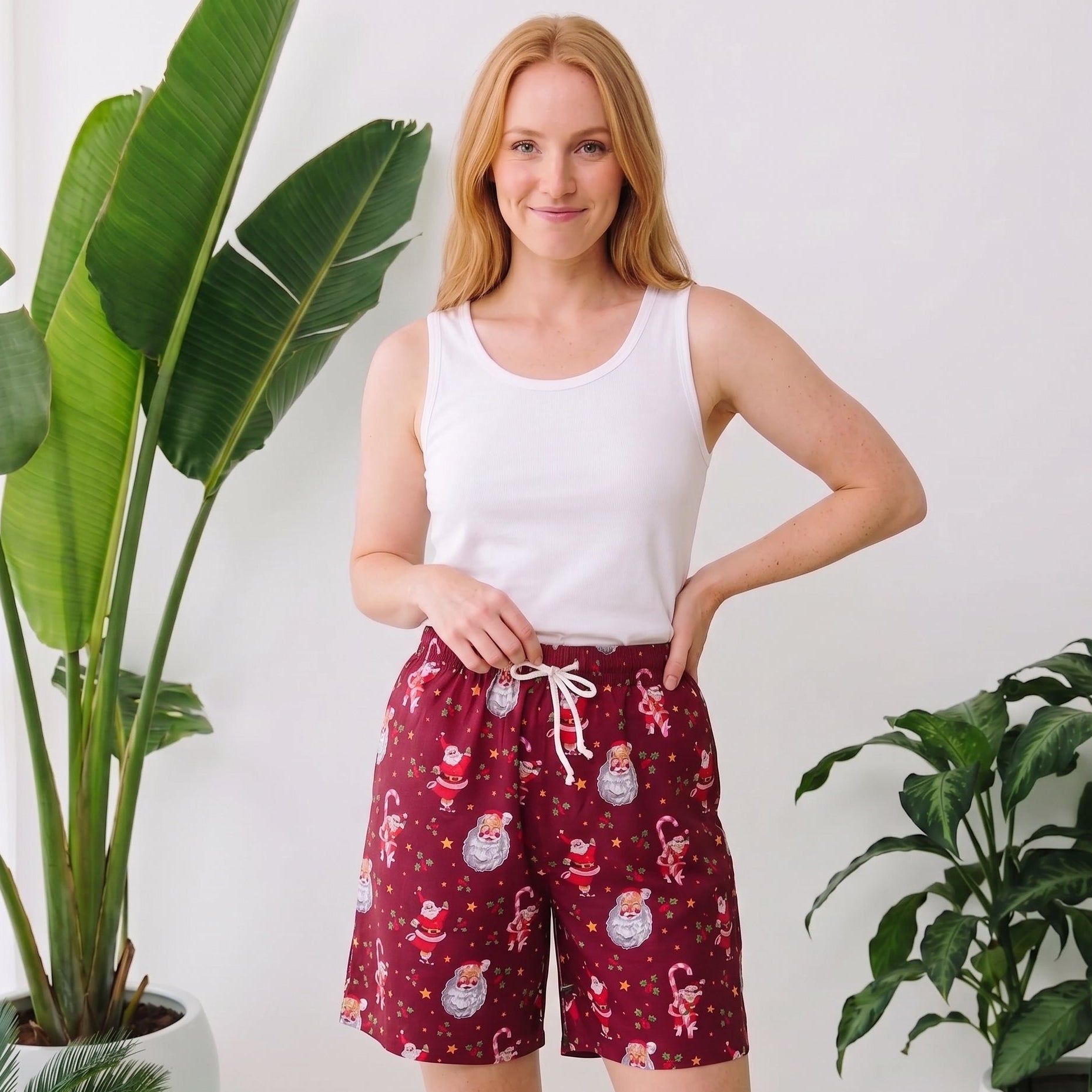A woman stands indoors wearing a white tank top and red holiday print shorts with a white drawstring. Large green potted plants are visible on both sides against a white wall.