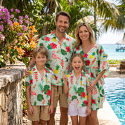 A family of four wearing matching tropical-print short-sleeve shirts and shorts stands together by a resort pool with palm trees and ocean in the background.