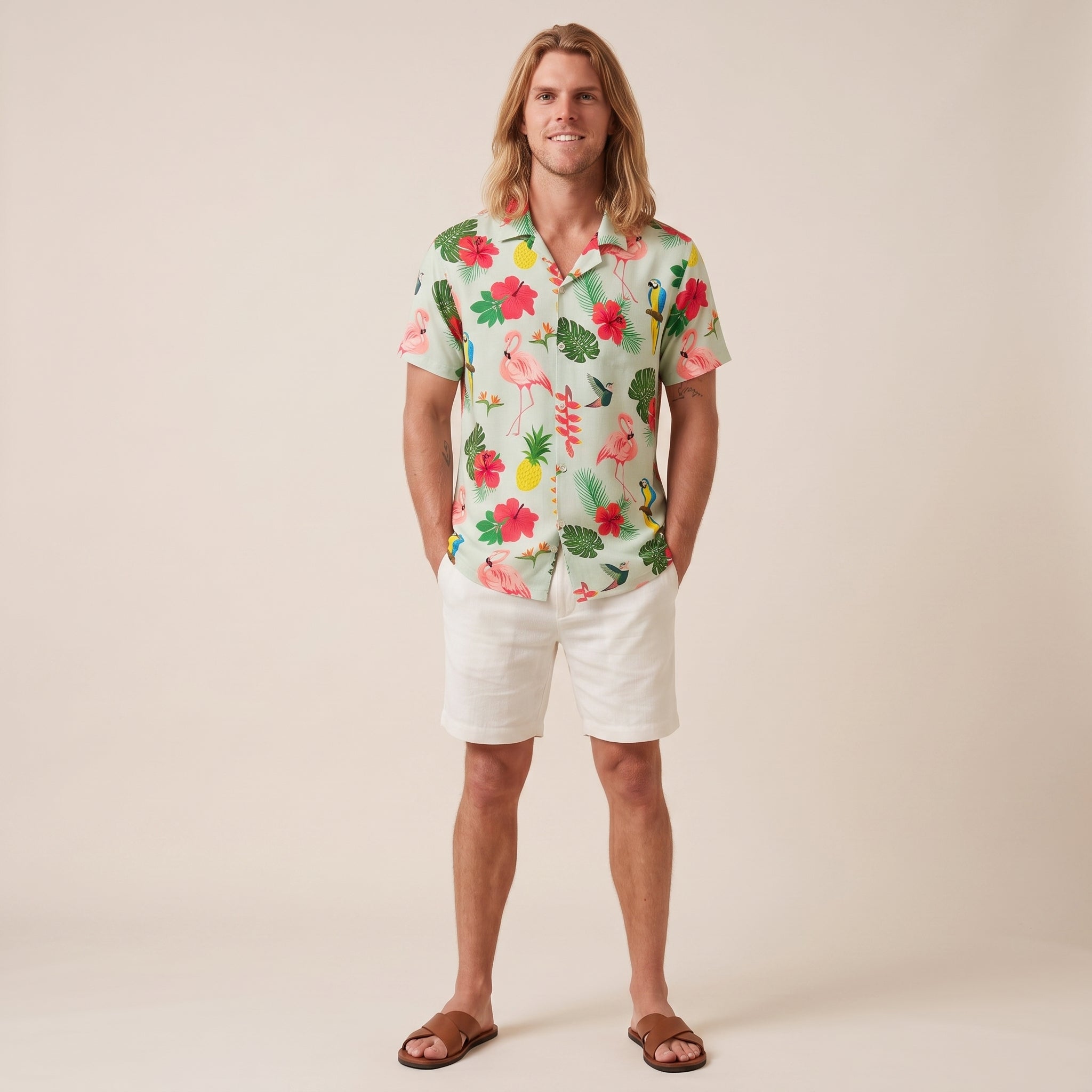 A man stands against a plain beige studio background wearing a short-sleeve tropical print button-up shirt with white shorts and brown sandals. The shirt features a light mint green base with pink flamingos, red hibiscus flowers, green leaves, and yellow pineapples.